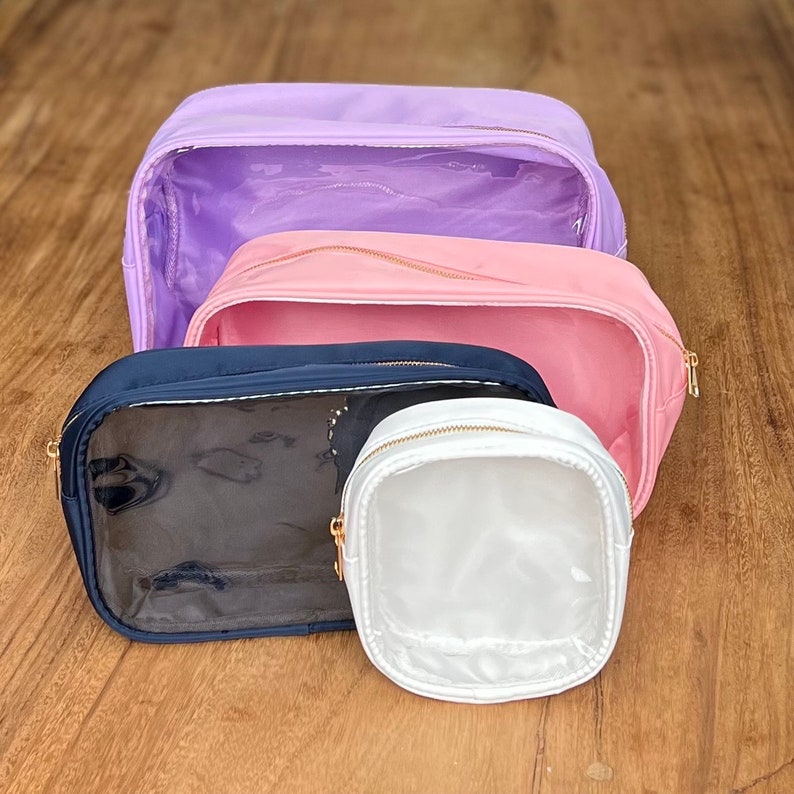 May include: Four clear makeup bags with gold zippers. The bags are in different sizes and colors: lavender, pink, navy blue, and white.