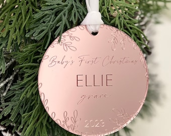 Puede incluir: Un adorno navideño de oro rosa con una cinta blanca. El adorno está decorado con un diseño floral y el texto "Baby's First Christmas Ellie grace 2023".