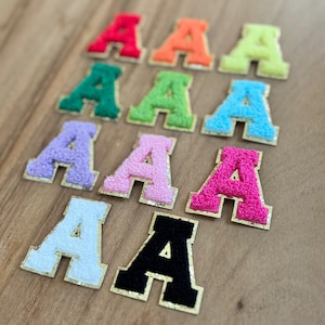 May include: A selection of fluffy letter "A" patches in a variety of colours. The patches include red, orange, green, blue, pink, purple, white, and black. Each letter has a gold border.