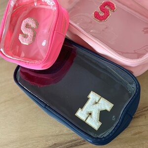 May include: Three clear zippered pouches with different colored fabric exteriors. The pouches have a clear plastic window and feature a large felt letter on the front. The pouches are pink, navy blue, and pink. The letters are S, K, and S.