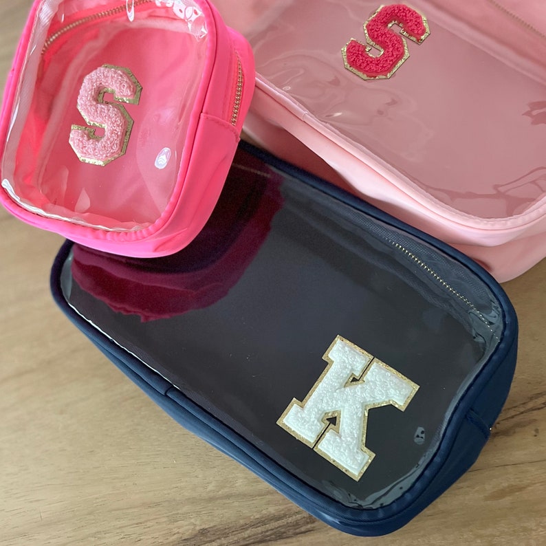 May include: Three clear zippered pouches with different colored fabric exteriors. The pouches have a clear plastic window and feature a large felt letter on the front. The pouches are pink, navy blue, and pink. The letters are S, K, and S.