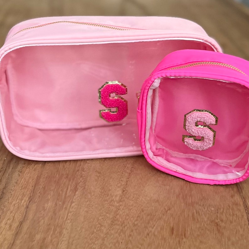 May include: Two pink clear makeup bags with gold zippers. The larger bag has a pink fluffy letter "S" on the front. The smaller bag has a pink fluffy letter "S" on the front.