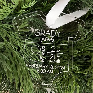 May include: Clear acrylic baby onesie ornament with white text. The ornament reads "GRADY james" and includes birth details: 8 pounds, 2 ounces, 54.6 cm, February 18, 2024, 5:30 AM. A white ribbon is attached for hanging.