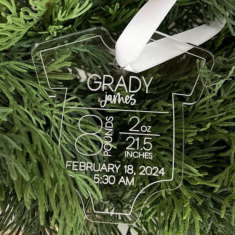 May include: Clear acrylic baby onesie ornament with white text. The ornament reads "GRADY james" and includes birth details: 8 pounds, 2 ounces, 54.6 cm, February 18, 2024, 5:30 AM. A white ribbon is attached for hanging.