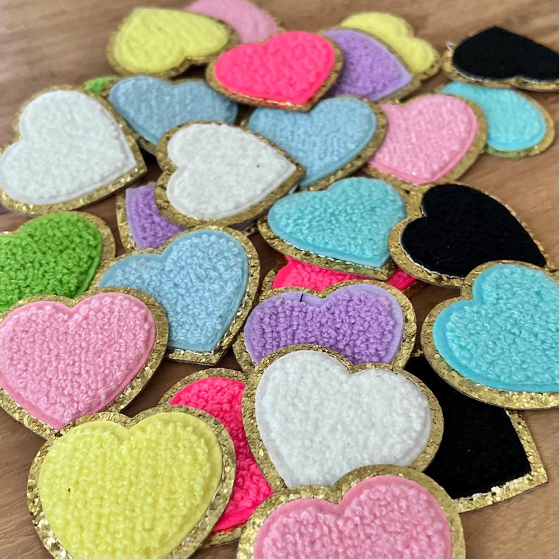 May include: A collection of colorful heart-shaped patches with a textured, fuzzy appearance. The patches come in various colors, including pink, yellow, blue, green, purple, white, and black, each outlined with a gold trim. These patches are suitable for crafts and embellishments.