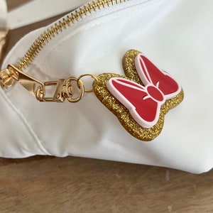 May include: A white fanny pack with a gold zipper and a gold chain. The chain has a red and pink bow charm with gold glitter.