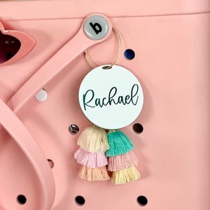 May include: White round tag with the name "Rachael" written in black cursive script. The tag is attached to a gold key ring with a pink and green tassel.