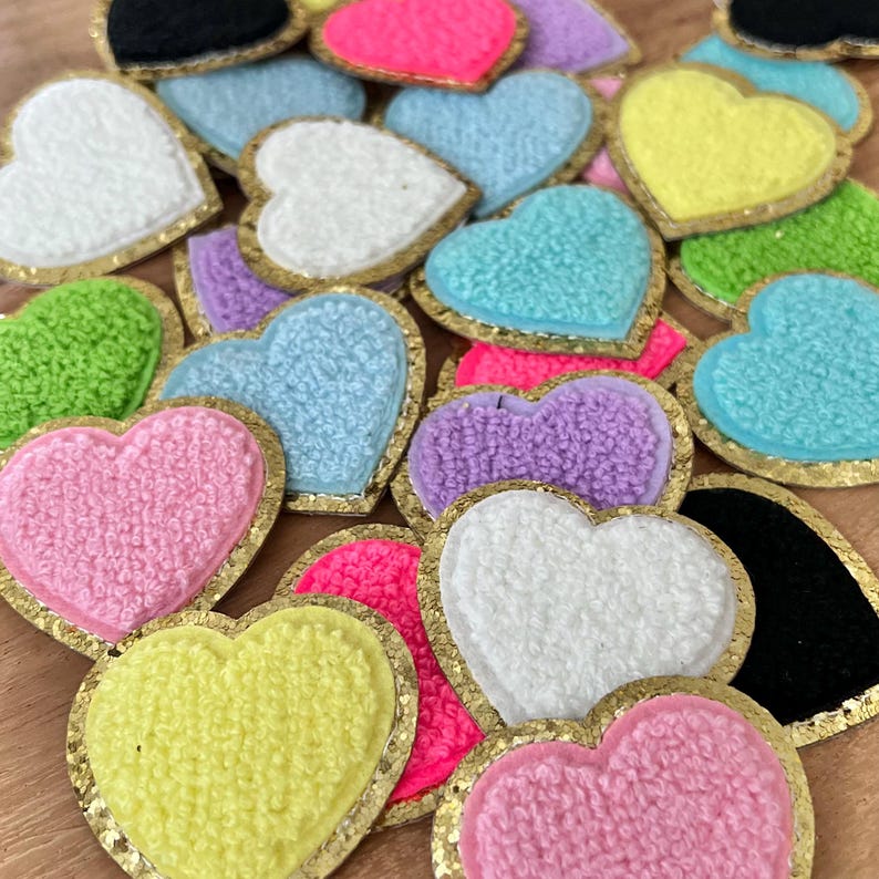 May include: A collection of colorful heart-shaped patches. Each patch features a textured, plush heart in various colors, including pink, blue, yellow, and green, outlined with a gold glitter border. The patches are scattered, creating a vibrant and playful display.
