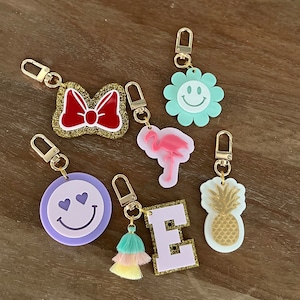 May include: A set of seven colorful keychains with different designs. The keychains include a bow, a flower, a flamingo, a pineapple, a smiley face, a letter E, and a tassel.