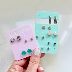 May include: A collection of stud earrings displayed on pink and teal earring holders. The earrings include various styles, such as small hoops, crystal studs, and gemstone spheres. The earring holders are held by a person with a white manicure.