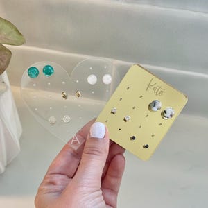 May include: A clear acrylic heart-shaped earring holder with various stud earrings. A gold rectangular earring holder with the name "Kate" is also visible. The earrings are in various colors and designs, including clear, gold, and teal.