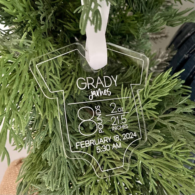 May include: Clear acrylic baby onesie ornament with white text. The ornament reads "GRADY james" with birth details: 3.6 kg, 57.2 cm, February 18, 2024, 5:30 AM. A white ribbon is attached for hanging.