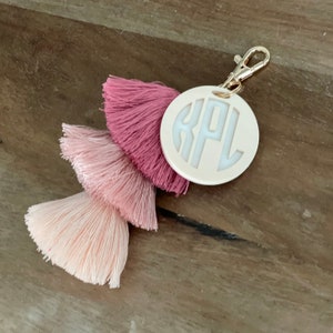 May include: A white circular keychain with the monogram "KPL" in white lettering. The keychain is attached to a pink, light pink, and white tassel.