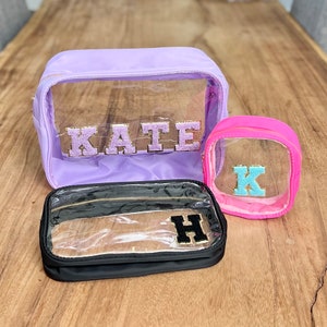 May include: Three clear cosmetic bags with different colored fabric exteriors. The largest bag is lavender and has the name "KATE" in gold glitter letters. The medium bag is black and has the letter "H" in gold glitter letters. The smallest bag is pink and has the letter "K" in blue glitter letters.