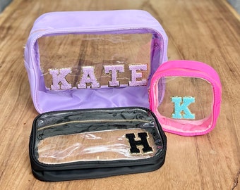 May include: Three clear cosmetic bags with different colored fabric exteriors. The largest bag is lavender and has the name "KATE" in gold glitter letters. The medium bag is black and has the letter "H" in gold glitter letters. The smallest bag is pink and has the letter "K" in blue glitter letters.