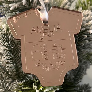 May include: Rose gold baby onesie ornament with the name "Amelia Grace" engraved. It includes the baby's birth weight of 2.72 kg, 113 grams, and length of 49.5 cm. The birth date and time are also engraved.