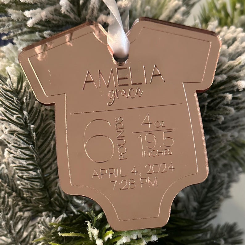 May include: Rose gold baby onesie ornament with the name "Amelia Grace" engraved. It includes the baby's birth weight of 2.72 kg, 113 grams, and length of 49.5 cm. The birth date and time are also engraved.