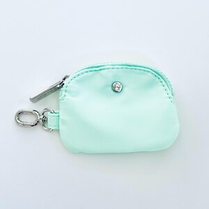 Puede incluir: Una peque&ntilde;a bolsa de cremallera de color verde claro con un tirador de cremallera plateado y un cierre de presi&oacute;n plateado. La bolsa est&aacute; unida a un llavero plateado.