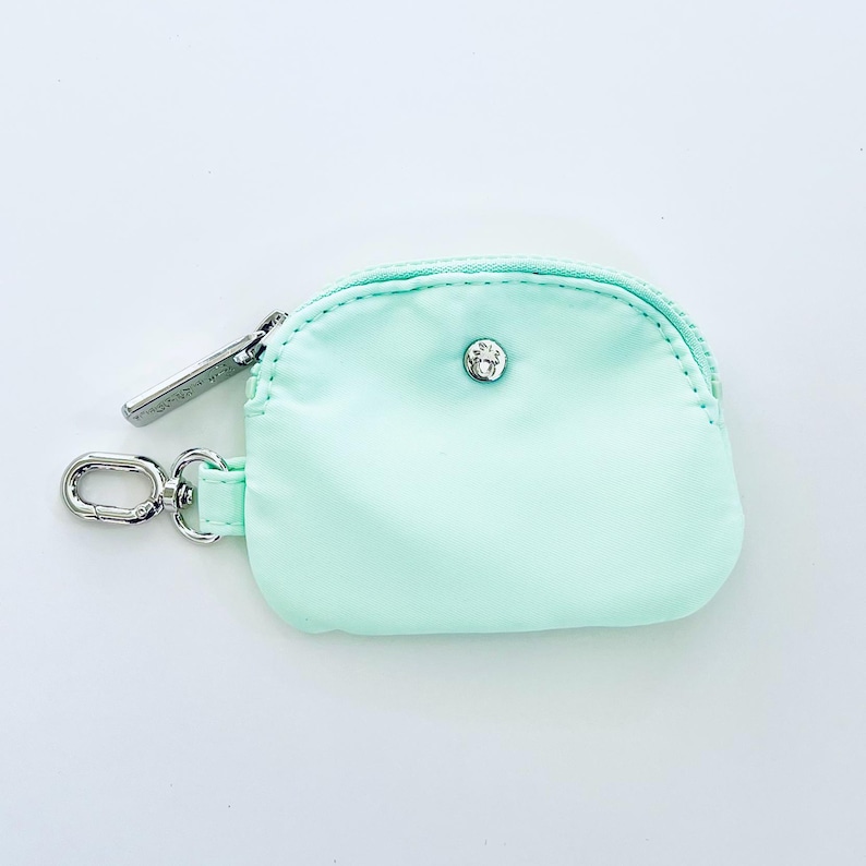 Puede incluir: Una peque&ntilde;a bolsa de cremallera de color verde claro con un tirador de cremallera plateado y un cierre de presi&oacute;n plateado. La bolsa est&aacute; unida a un llavero plateado.