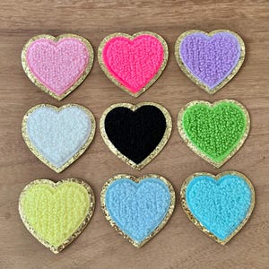 May include: Nine heart-shaped patches in various colors, including pink, black, and green. Each patch has a textured center and a gold glitter border. These patches are suitable for crafts and embellishments.