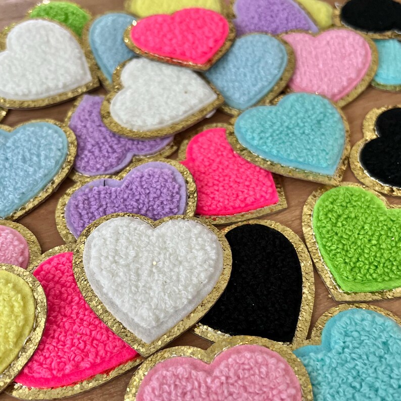 May include: A collection of colorful heart-shaped patches. The patches feature a textured, fuzzy surface in various colors, including white, pink, purple, blue, and green. Each heart is outlined with a gold metallic trim, creating a decorative effect.