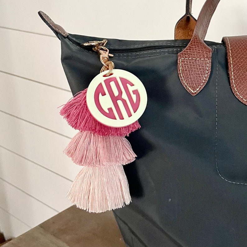 May include: A white circle keychain with the monogram "CRG" in pink letters. The keychain is attached to a dark gray tote bag with a brown leather handle. The keychain also has a pink, white, and light pink tassel.