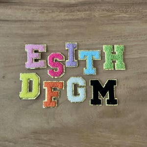 May include: A selection of embroidered fabric letters in a variety of colours, such as pink, orange, and green. Each letter has a gold border and a fuzzy texture, suitable for crafting or customisation.