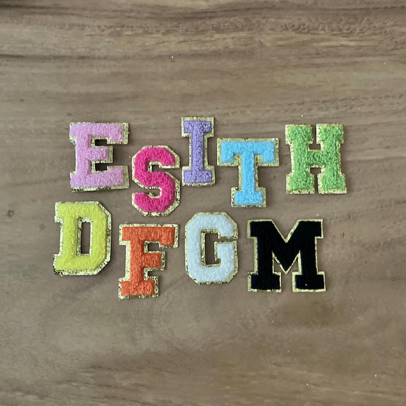 May include: A selection of embroidered fabric letters in a variety of colours, such as pink, orange, and green. Each letter has a gold border and a fuzzy texture, suitable for crafting or customisation.