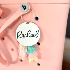 May include: White round tag with the name "Rachael" written in black cursive lettering. The tag is attached to a pink tote bag with a tassel.