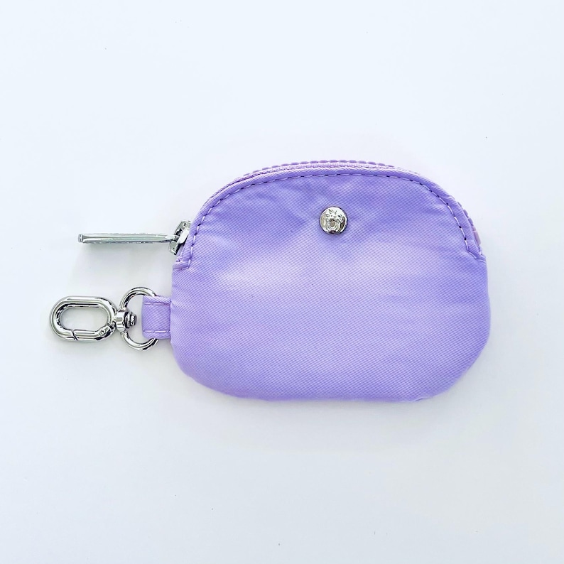 Puede incluir: Una peque&ntilde;a cartera de monedas de color lavanda con una cremallera plateada y un cierre de bot&oacute;n plateado. La cartera tiene un llavero plateado.