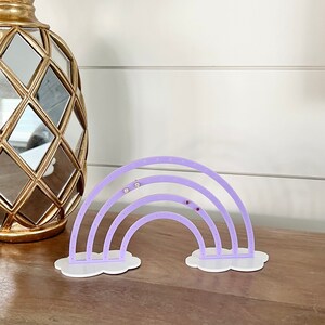 May include: A lavender acrylic earring holder shaped like a rainbow, with white cloud-shaped bases. The holder has multiple holes for displaying earrings. A gold decorative object is in the background.