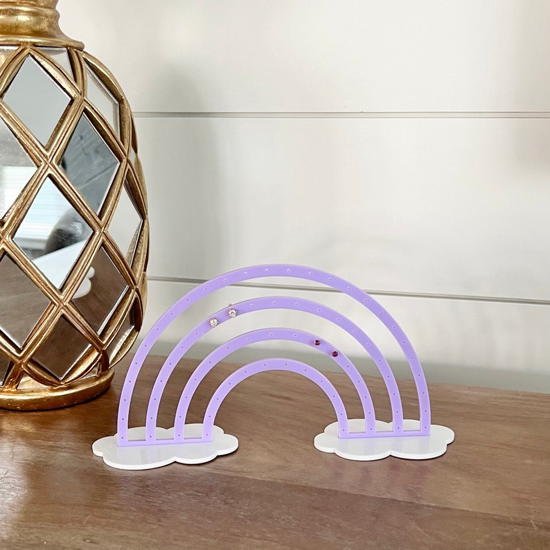 May include: A lavender acrylic earring holder shaped like a rainbow, with white cloud-shaped bases. The holder has multiple holes for displaying earrings. A gold decorative object is in the background.