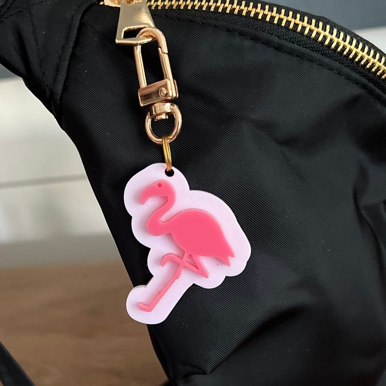 May include: A pink flamingo keychain charm attached to a gold key ring. The charm is made of plastic and has a white outline.