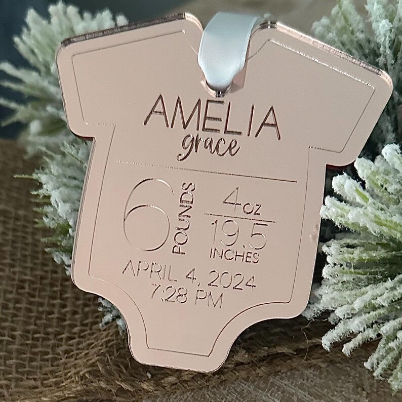May include: A rose gold babygrow-shaped keepsake engraved with "AMELIA grace". It shows birth details: 6 pounds, 4 ounces, 19.5 inches (49.5 cm), April 4, 2024, 7:28 PM. A white ribbon is attached for hanging.