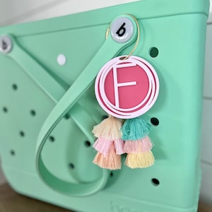 May include: A light green tote bag with the word "bogg" printed on the bottom. The bag has a white circle with a black "b" on it and a pink circle with a white "F" on it attached to the handle. The bag has a multi-colored tassel attached to the pink circle.