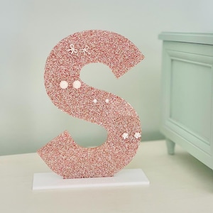 May include: A large, glitter-covered letter "S" in a rose-gold hue, standing on a white base. The letter is adorned with small, white decorative accents, including bows and pearls. The background features a light blue wall and a mint-green cabinet.