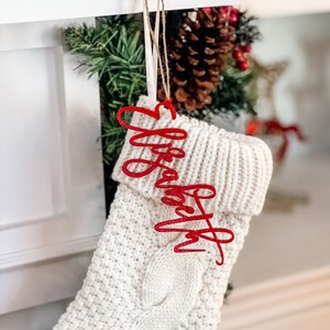 May include: A cream-colored cable knit Christmas stocking featuring a red glittery acrylic name tag that reads Elizabeth in a cursive script font. The tag is attached to the stocking with a thin twine loop, hanging from the top cuff.