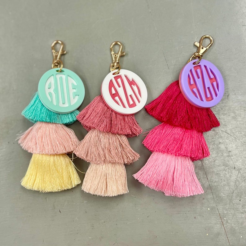 May include: Three tassel keychains with personalized monogrammed circles. The circles are in mint green, white, and purple with white lettering. The tassels are layered in shades of pink and yellow.