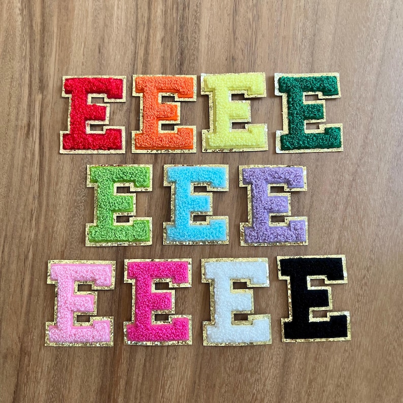 May include: A selection of embroidered letter "E" patches in various colours. Each letter is a different colour, such as red, orange, yellow, green, blue, purple, pink, white, and black, with a gold border. These patches are likely for clothing or craft projects.