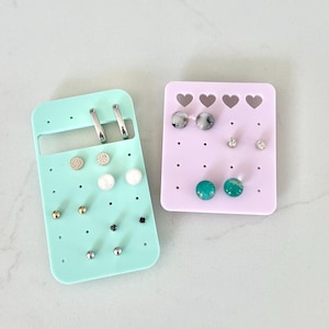 Peut inclure: Deux présentoirs à boucles d'oreilles, un vert menthe et un lavande. Le support vert menthe a une forme rectangulaire avec une poignée et contient diverses boucles d'oreilles et créoles. Le support lavande a un dessus en forme de cœur.