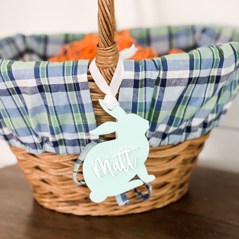 May include: A light blue bunny-shaped Easter basket tag with the name "Matt" written in white. The tag is hanging from a wicker basket lined with a blue and green plaid fabric.