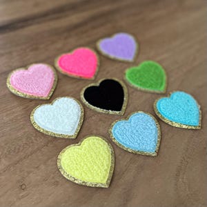 May include: A collection of colorful heart-shaped patches. Each patch features a textured surface and a gold-colored border. The hearts come in various colors, including pink, neon pink, purple, green, white, black, light blue, and yellow.