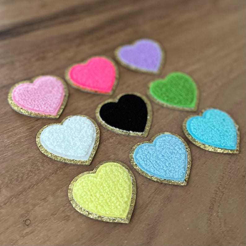 May include: A collection of colorful heart-shaped patches. Each patch features a textured surface and a gold-colored border. The hearts come in various colors, including pink, neon pink, purple, green, white, black, light blue, and yellow.