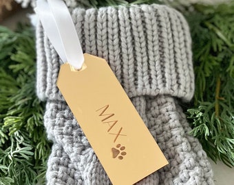May include: A light grey knitted stocking featuring a gold-coloured rectangular acrylic tag attached with a white ribbon. The tag is engraved with the name MAX and a paw print icon, positioned against a background of green pine needles.
