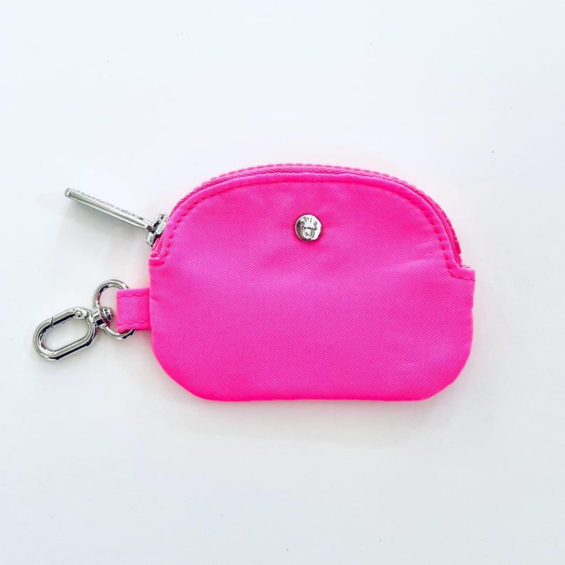 Puede incluir: Una peque&ntilde;a bolsa rosa brillante con cremallera y un tirador de cremallera plateado y un cierre de bot&oacute;n plateado. La bolsa tiene un llavero.