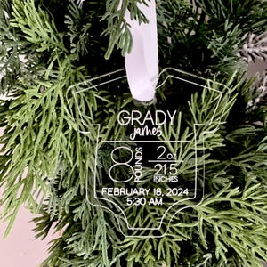 May include: Clear acrylic baby onesie ornament with the name "Grady James" and birth details: 8 pounds, 2 ounces, 54.6 cm, born February 18, 2024, at 5:30 AM. The ornament hangs from a white ribbon, set against a backdrop of green foliage.