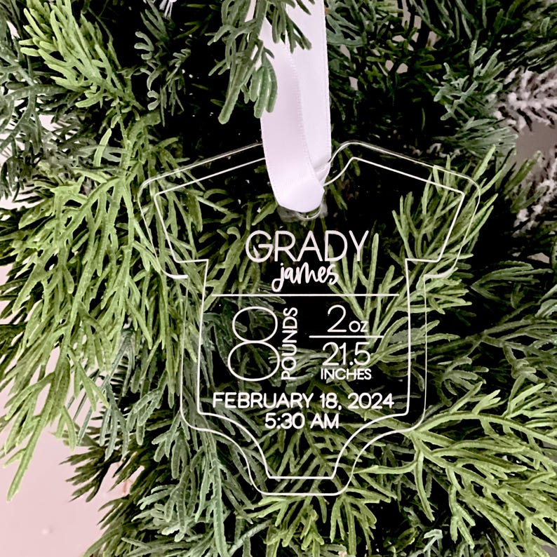 May include: Clear acrylic baby onesie ornament with the name "Grady James" and birth details: 8 pounds, 2 ounces, 54.6 cm, born February 18, 2024, at 5:30 AM. The ornament hangs from a white ribbon, set against a backdrop of green foliage.