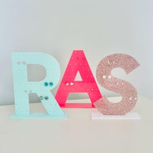 May include: Earring display stands in the shape of the letters R, A, and S. The R is light blue, the A is pink, and the S is glittery rose gold. Each letter has holes for displaying earrings.