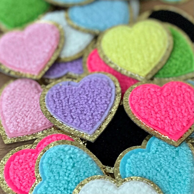 May include: A collection of colorful heart-shaped patches. The patches feature a textured, fuzzy surface in various shades including pink, purple, yellow, and blue. Each heart is outlined with a gold trim.