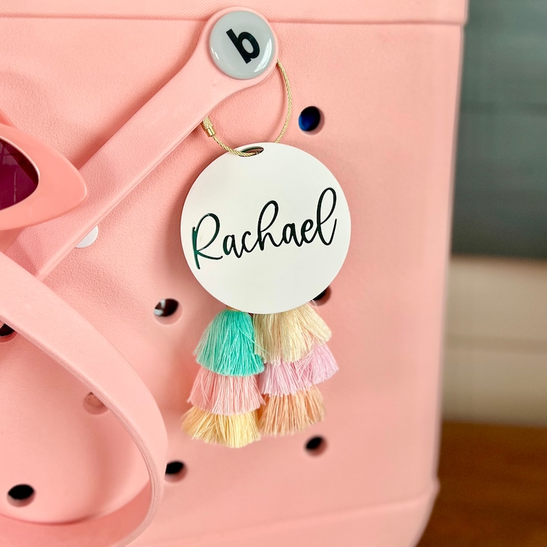 May include: White round tag with the name "Rachael" written in black cursive script. The tag is attached to a pink bag with a gold ring and colorful tassels.